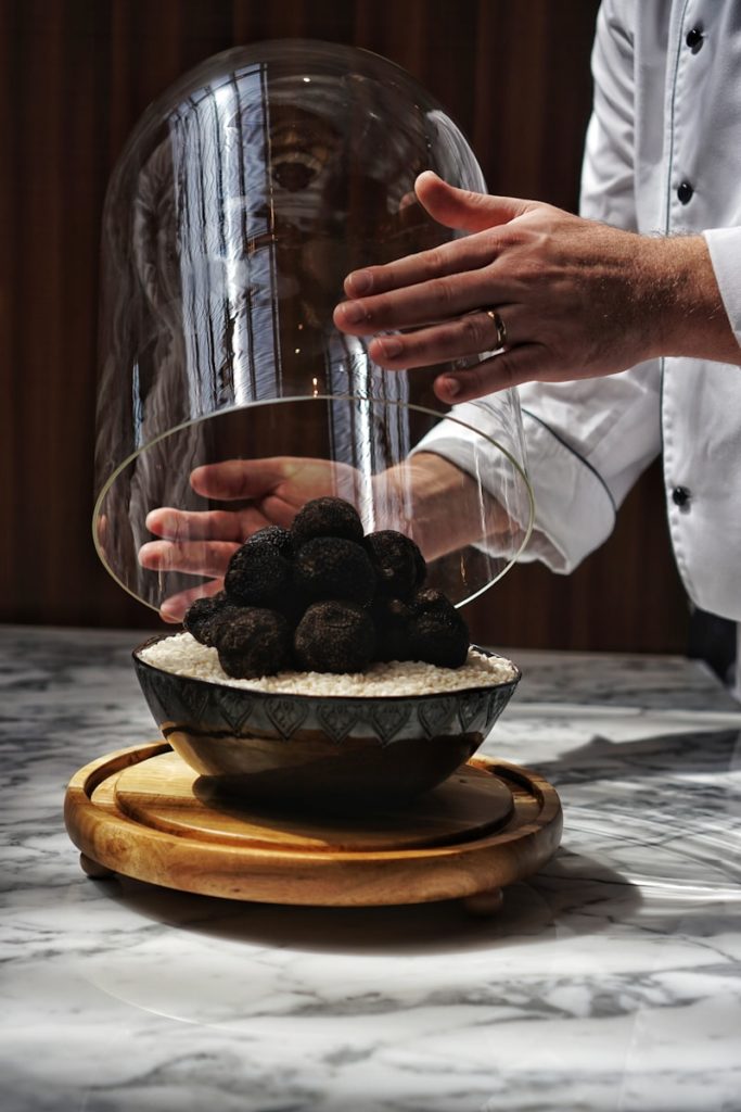 Chef cuisinier soulevant une large coupole en verre pour révéler des truffes noires disposées avec soin.